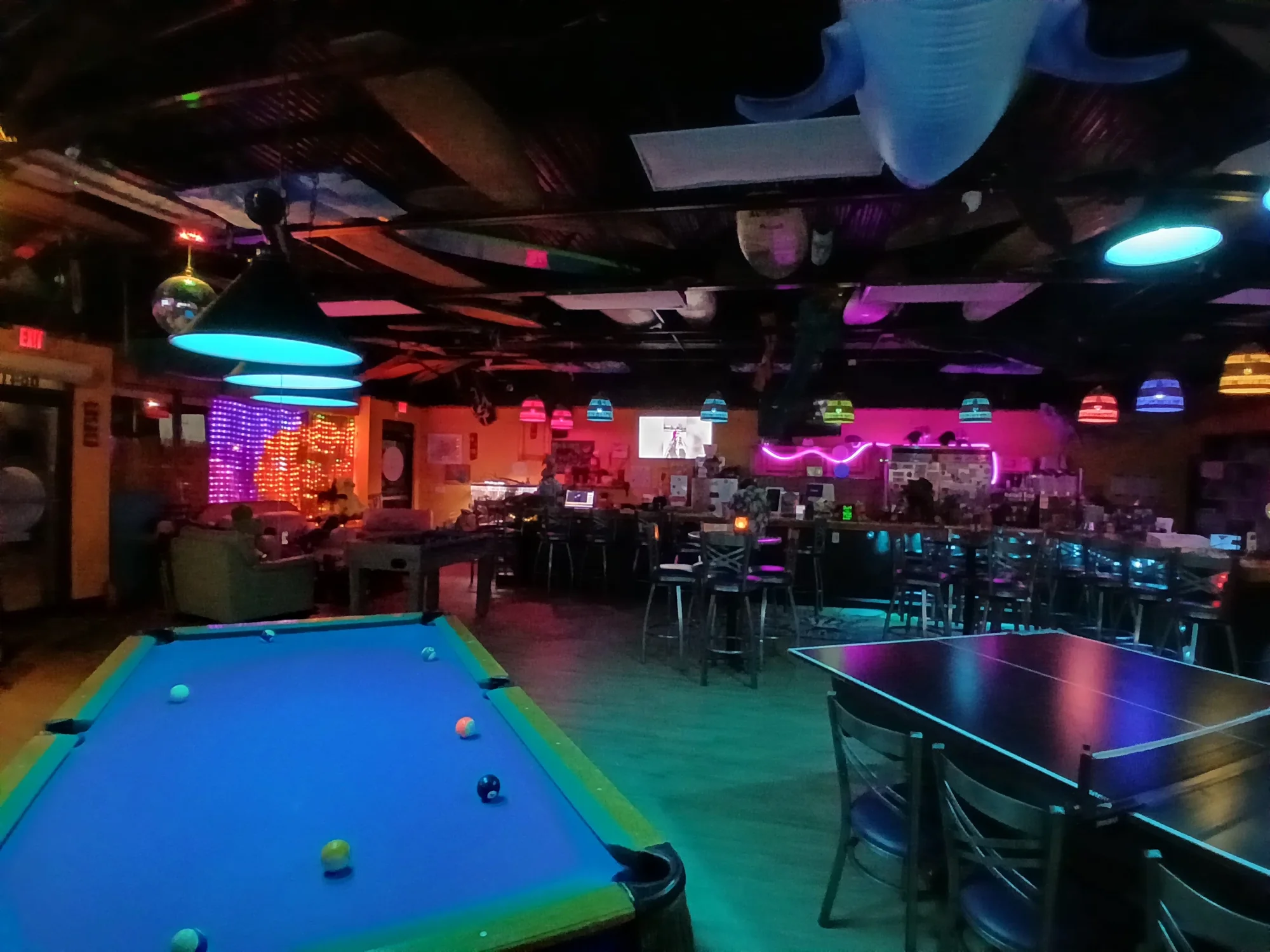 The main room at night: a blue-felt pool table under neon light, surfboards hung from the ceiling and the bar glowing in the back